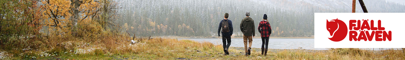 Three people are walking towards a lake, surrounded by mountains, wearing Fjallraven gear.