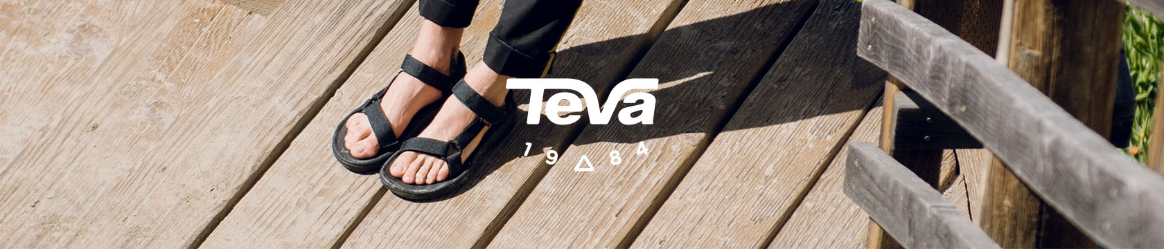 Person's feet standing on wooden stairs wearing Teva sandals.