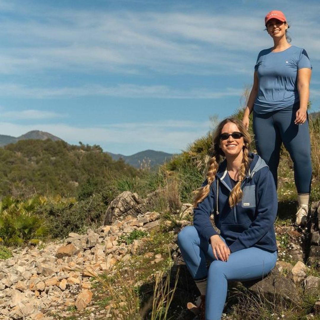 Two women looking sat on a rock wearing ACAI with mountains in the background