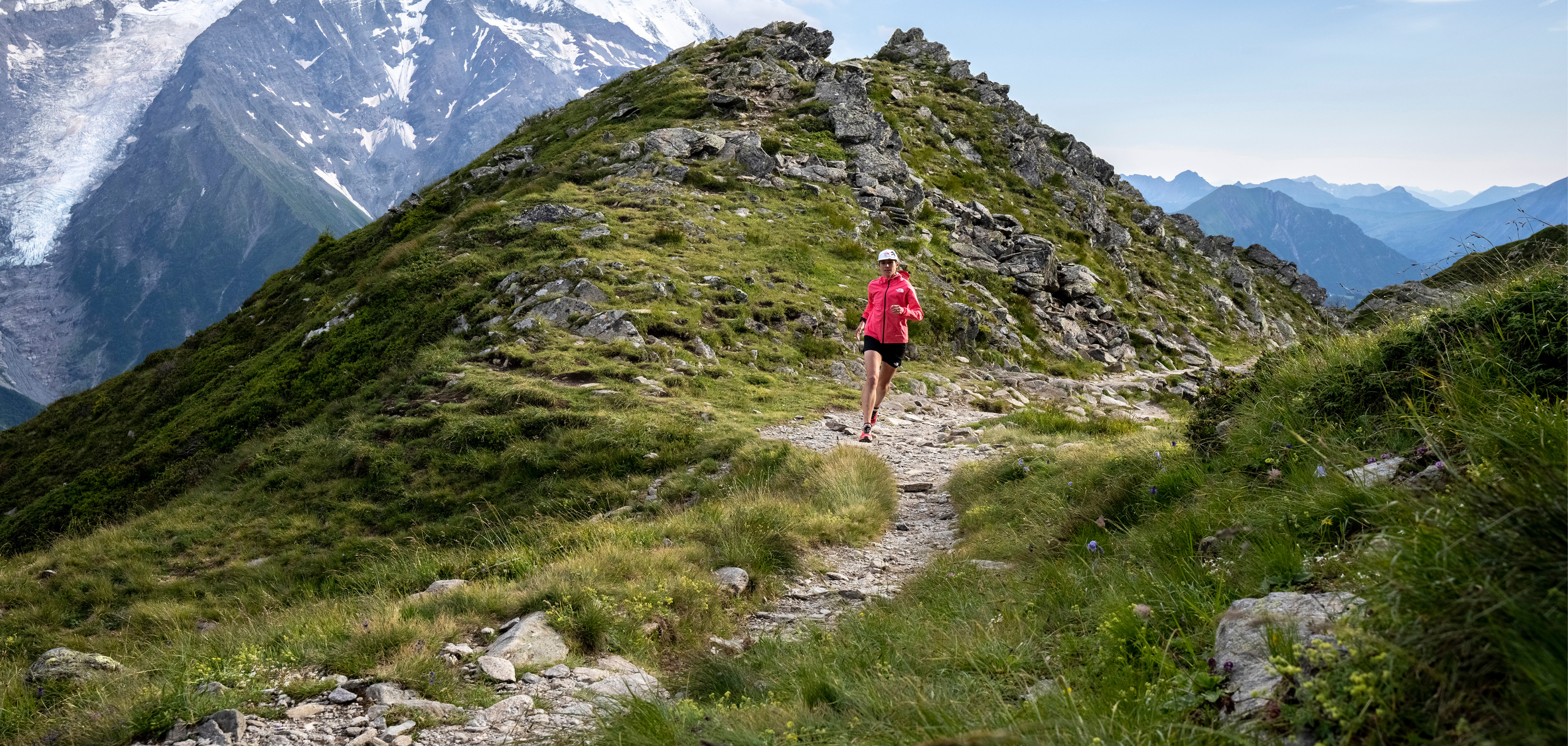 A woman in trail kit running along a trail with green grassy mountains in the background