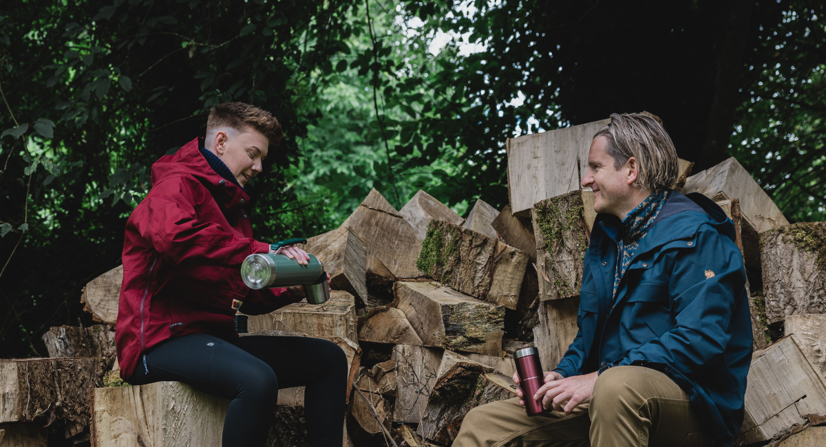 A woman in a bright red anorak laughs with a friend while pouring a hot drink from a green stanley thermos