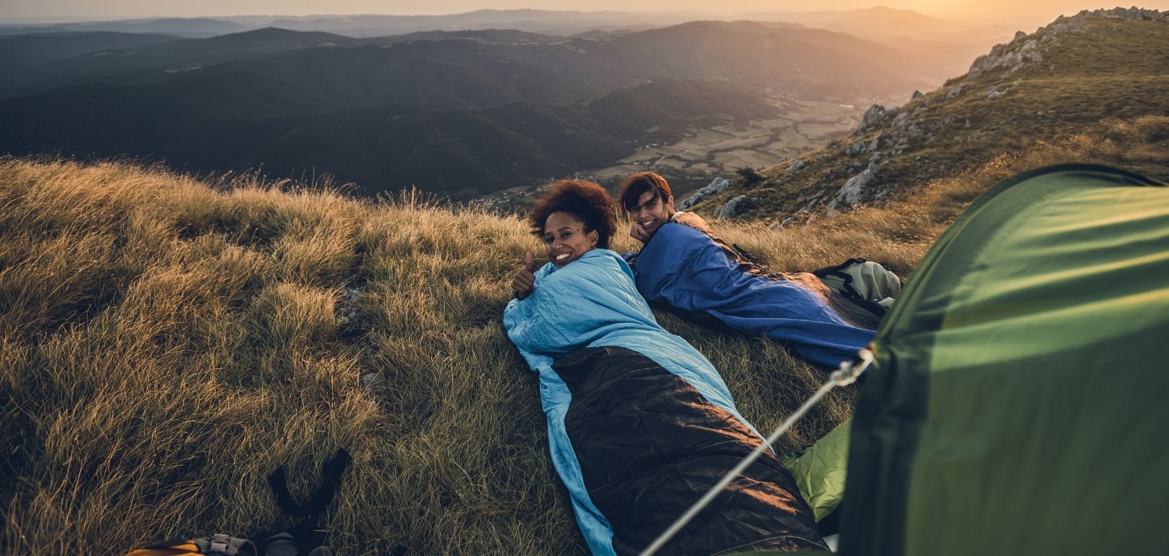 Two women look back at the camera laid in their sleeping bags in the grass beside their tent