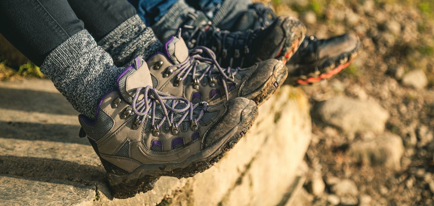 Two pairs of hiking boots worn by two people balance on the edge of a rock. The front most pair have vibrant purple laces
