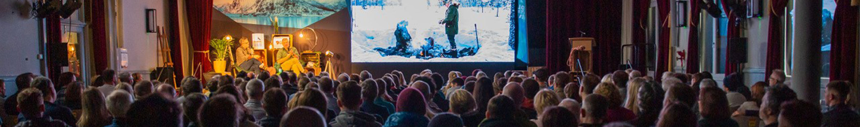 People are watching a video on a not big scree in a theatre-like setting.