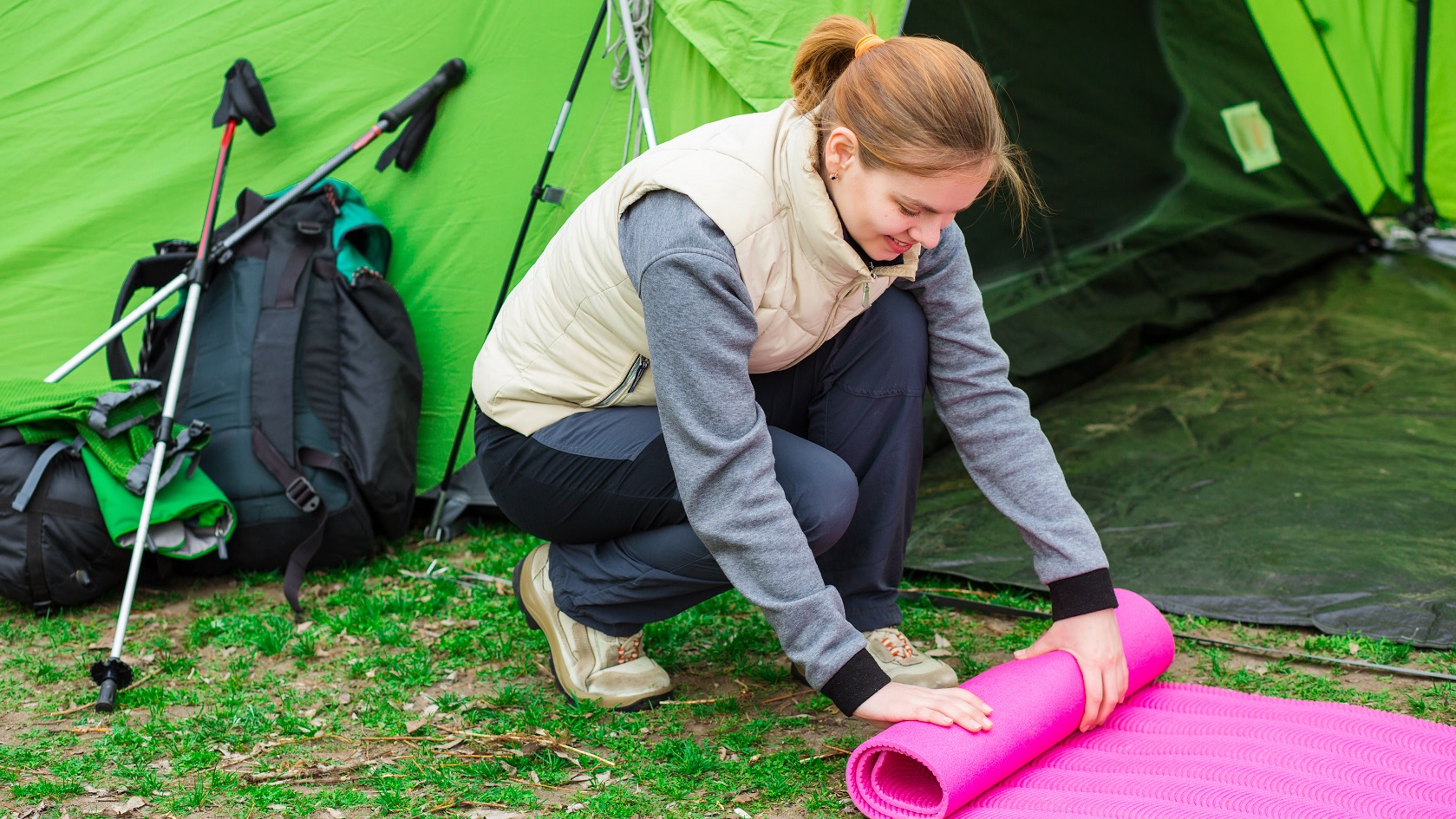 Cheerful woman rolling camping mat , during the camping holidays. In the background tourist tent and equipment for outdoor activities.