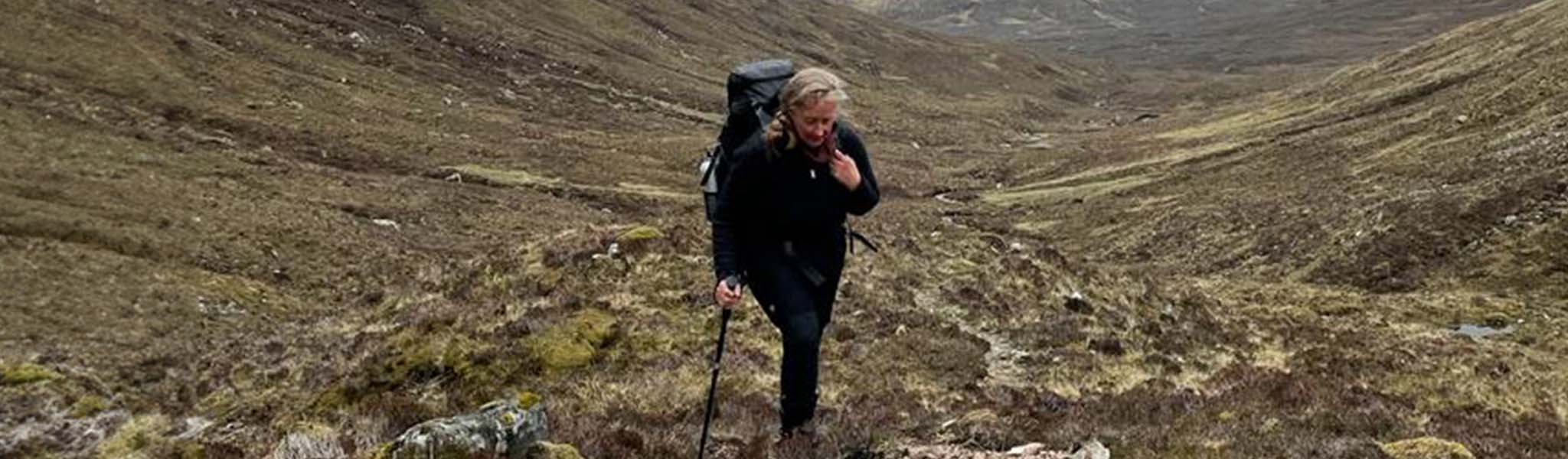 A woman is hiking, wearing walking kit and a rucksack, using a trekking pole.