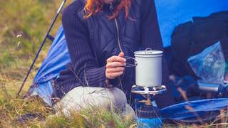 Young woman is sitting in her tent and cooking with a portable camping stove